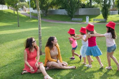 Happy children playing in a sunny park with two mothers sitting nearby, enjoying the outdoors.